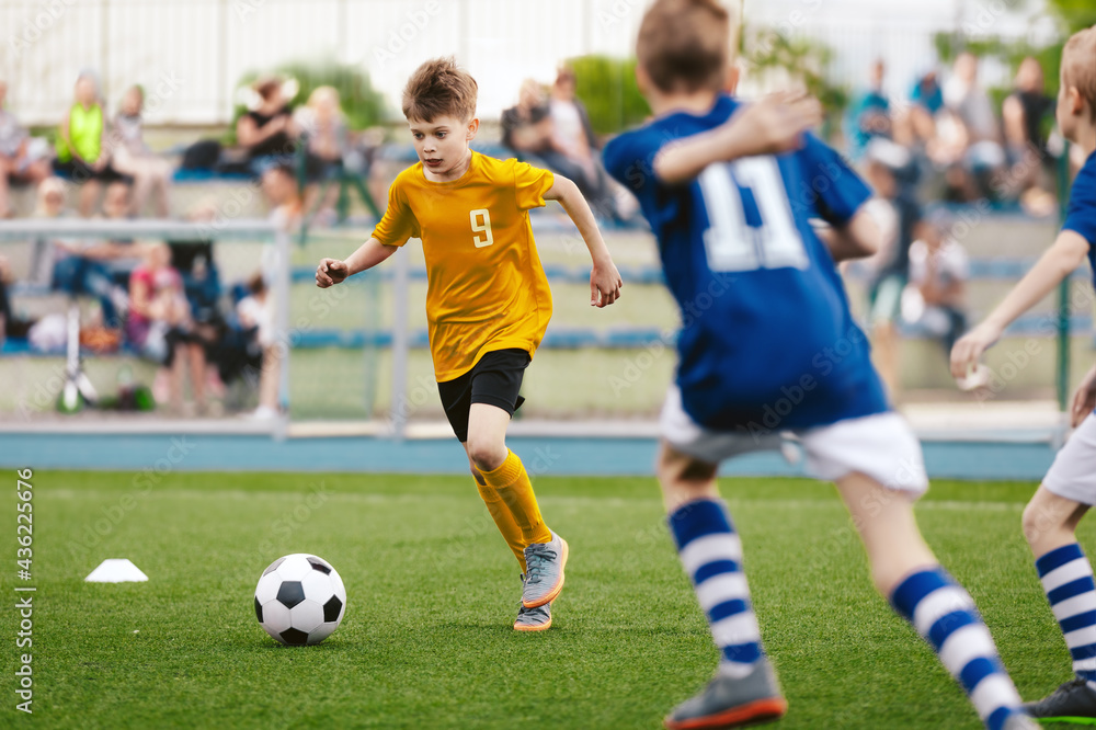 Young Boy Running Fast and Kicking Soccer Ball on Grass Pitch. Football Tournament For Youth School Teams. Group of Children in Jersey Unifrom Playing Sports. Football Fans and Parents in Background Young Boy Running Fast and Kicking Soccer Ball on Grass Pitch. Football Tournament For Youth School Teams. Group of Children in Jersey Unifrom Playing Sports. Football Fans and Parents in Background