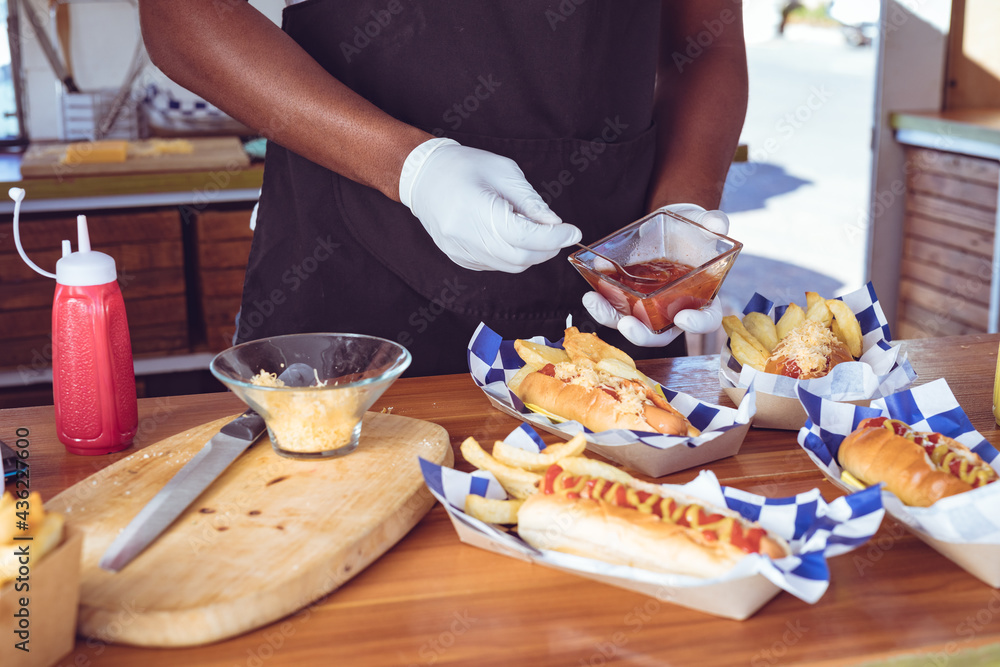 Midsection of african american man in food truck holding ketchup dish with hotdogs on worktop Midsection of african american man in food truck holding ketchup dish with hotdogs on worktop