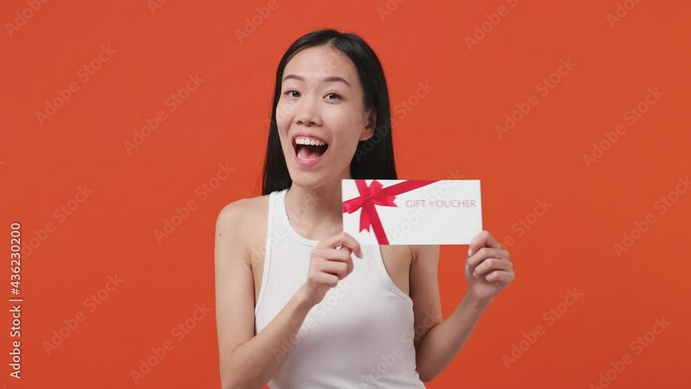 Surprised shocked happy young asian woman 20s years old wears white tank top shirt pointing finger on gift certificate voucher for store doing winner gesture isolated on orange color background studio Surprised shocked happy young asian woman 20s years old wears white tank top shirt pointing finger on gift certificate voucher for store doing winner gesture isolated on orange color background studio