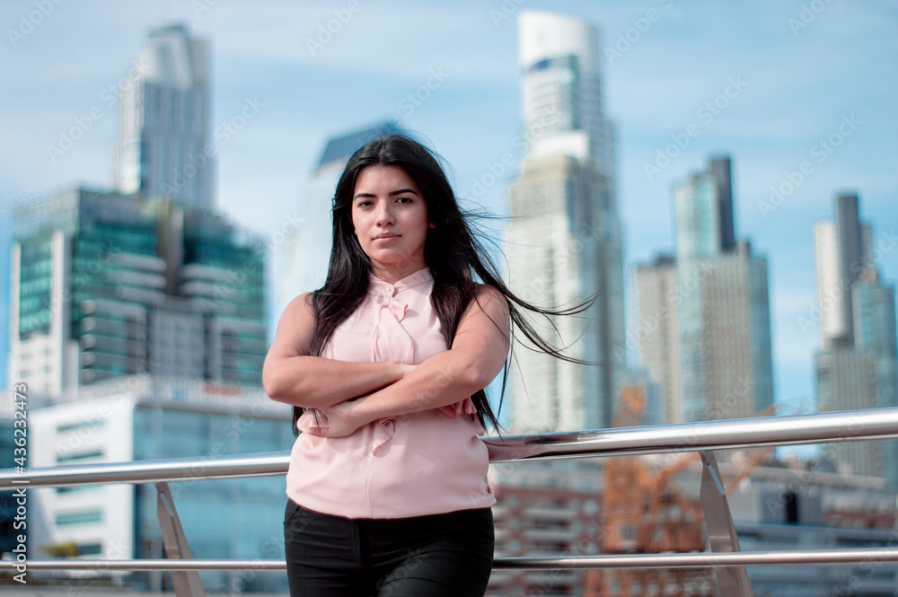 girl posing with her arms crossed leaning against a railing with business buildings and commercial offices all around her.
