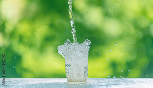Water flows into a glass placed on a wooden bar,over sunlight and natural green background.blur shadow.