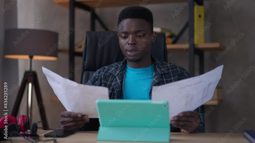 Front view portrait of busy young male analyst examining business graphs holding head in hands. Overburdened African American man sighing planning strategy in home office sitting at table