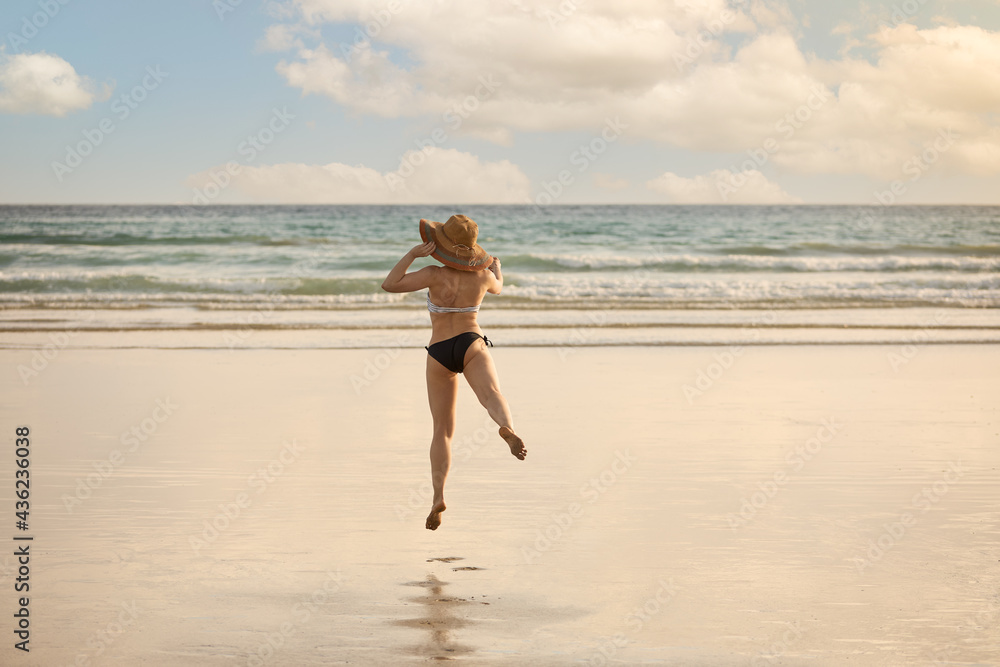 Girl in bikini with straw hat jumping on the shore of a beach.
