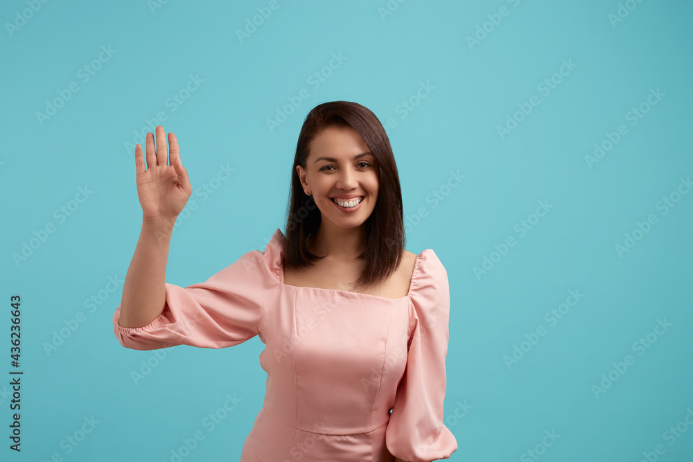 Hello guys. Satisfied beautiful woman in pink dress shows palm and welcomes everyone smiles positively, makes waving gesture, isolated over blue background. People expression concept. Hello guys. Satisfied beautiful woman in pink dress shows palm and welcomes everyone smiles positively, makes waving gesture, isolated over blue background. People expression concept.