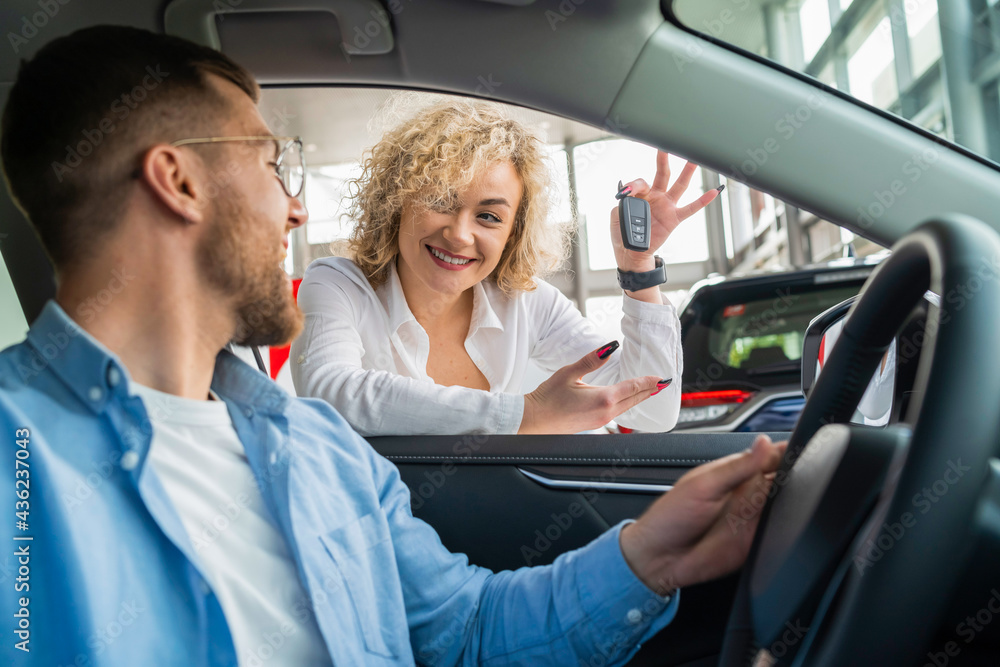 Young couple buying new car. Woman shows key to new auto to her husband who is sitting in car at dealership Young couple buying new car. Woman shows key to new auto to her husband who is sitting in car at dealership