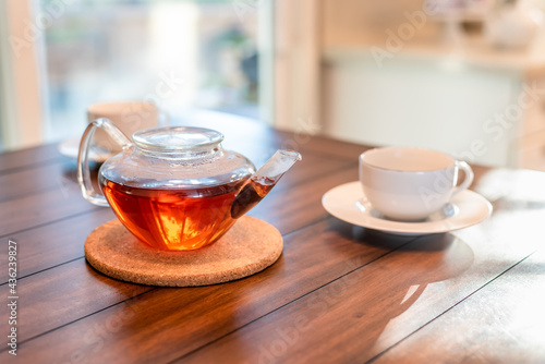 Delicious brewed tea in a glass teapot with a white porcelain cup at the ready on a rich wood grained table in a bright kitchen.