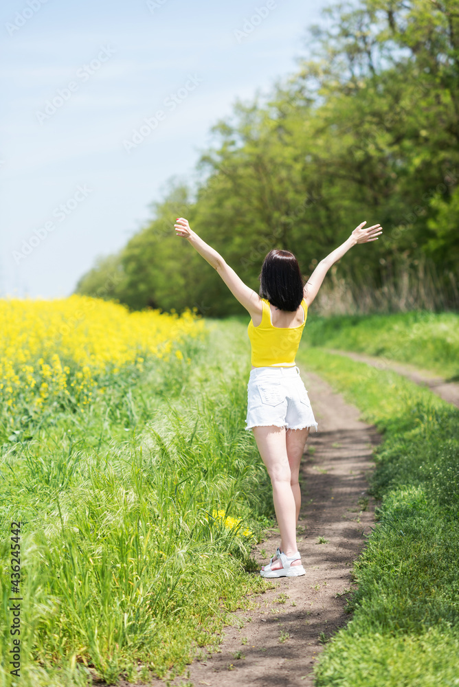 Young beautiful girl stands with her back to the camera and looks at the rapeseed yellow field, raising her hands up Young beautiful girl stands with her back to the camera and looks at the rapeseed yellow field, raising her hands up