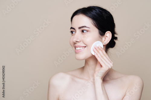 Beautiful Caucasian woman gently cleaning face with makeup remover pad ion beige background