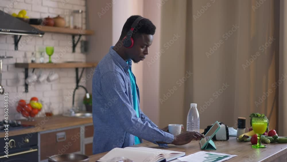 Side view of young concentrated man in headphones drinking morning coffee surfing Internet checking e-mail on tablet standing at kitchen countertop. Focused African American millennial in home office Side view of young concentrated man in headphones drinking morning coffee surfing Internet checking e-mail on tablet standing at kitchen countertop. Focused African American millennial in home office