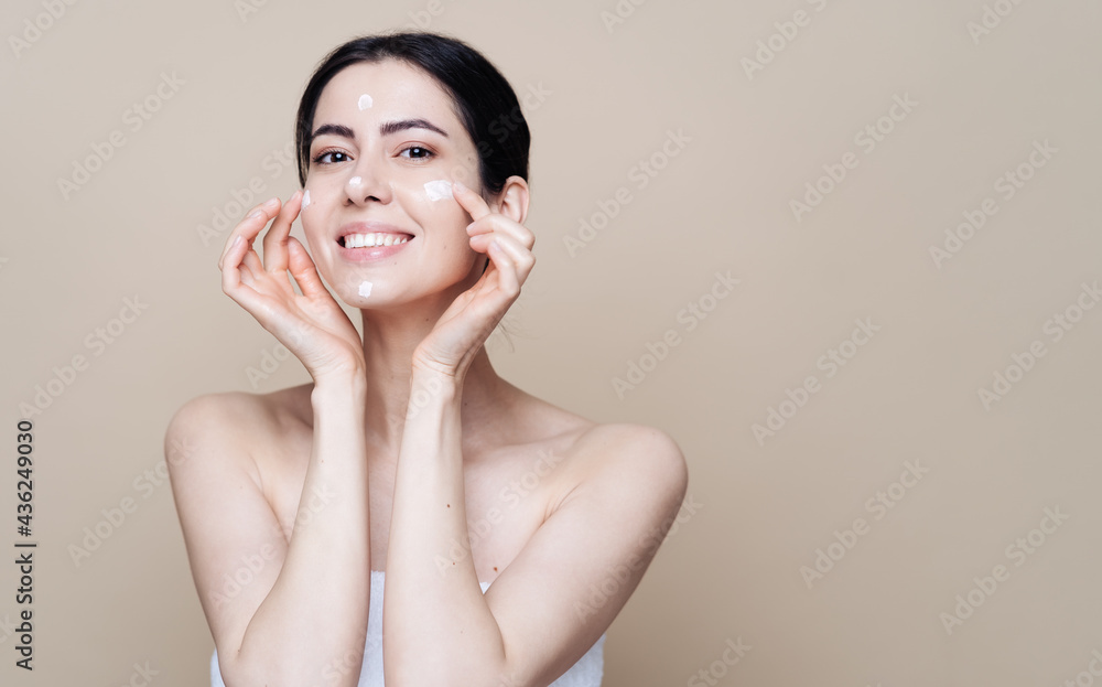 Close up beauty portrait of a laughing beautiful half naked woman applying face cream over beige background with copy space