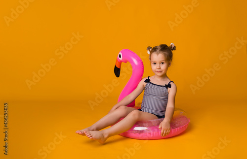 cute little child in a striped swimsuit sits in a pink inflatable circle and looks at the camera on a yellow background with space for text