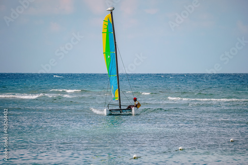 Scenic view at small catamaran with yellow green blue sail sailing in sea. An unrecognizable men enjoy alone tropical sea. Small waves on turquoise surface, clean blue skies