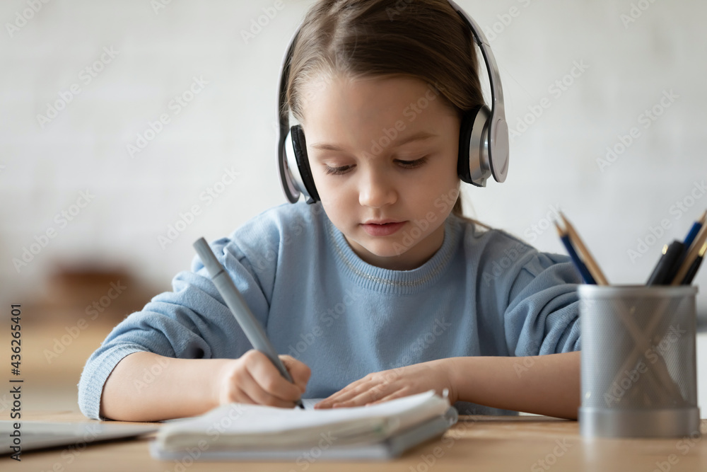 Close up of smart small schoolgirl in headphones write prepare homework alone at home. Little Caucasian 7s girl child in earphones handwrite study online distant, home assignment. Education concept.