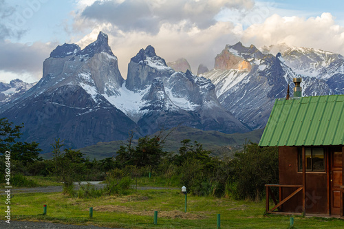 Chalé de madeira com telhado verde com gramado verde e arbustos ao redor e ao fundo a bela paisagem das montanhas Cuernos del Paine