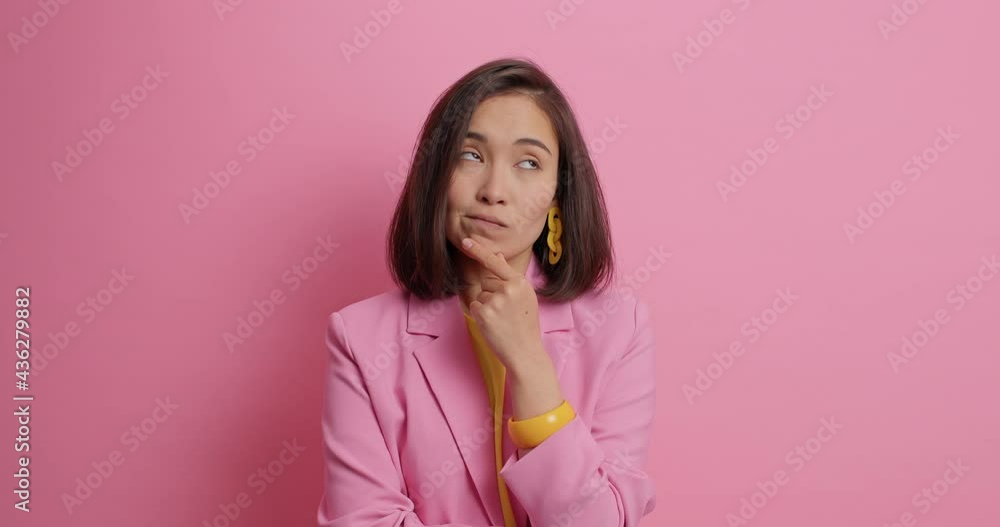 Stylish elegant woman in business costume holds chin looks around with thoughtful expression tries to decide then smiles gladfully has dark hair isolated over pink background. Let me think concept Stylish elegant woman in business costume holds chin looks around with thoughtful expression tries to decide then smiles gladfully has dark hair isolated over pink background. Let me think concept