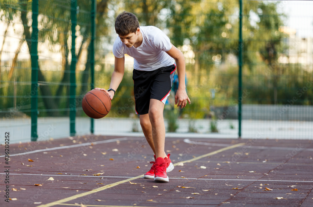 Obraz premium A cute boy in a white t-shirt plays basketball on a city playground. Active teen enjoying an outdoor game with an orange ball. Hobby, active lifestyle, sport for kids.