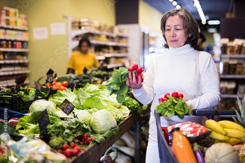 Portrait of positive elderly woman buying fresh organic vegetables in supermarket, choosing red radish © JackF