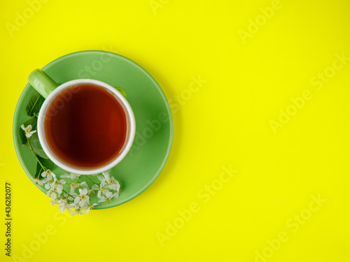 Green cup of black tea with white bird cherry tree flowers in saucer on bright illumitated yellow paper background. flat lay, top view, copy space, vertical horizontal