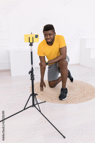 Focused sporty afro american sit on floor at home doung sport and watching tutorials online on smartphone . Millennial train at home having sport class on yellow phone after work  photo