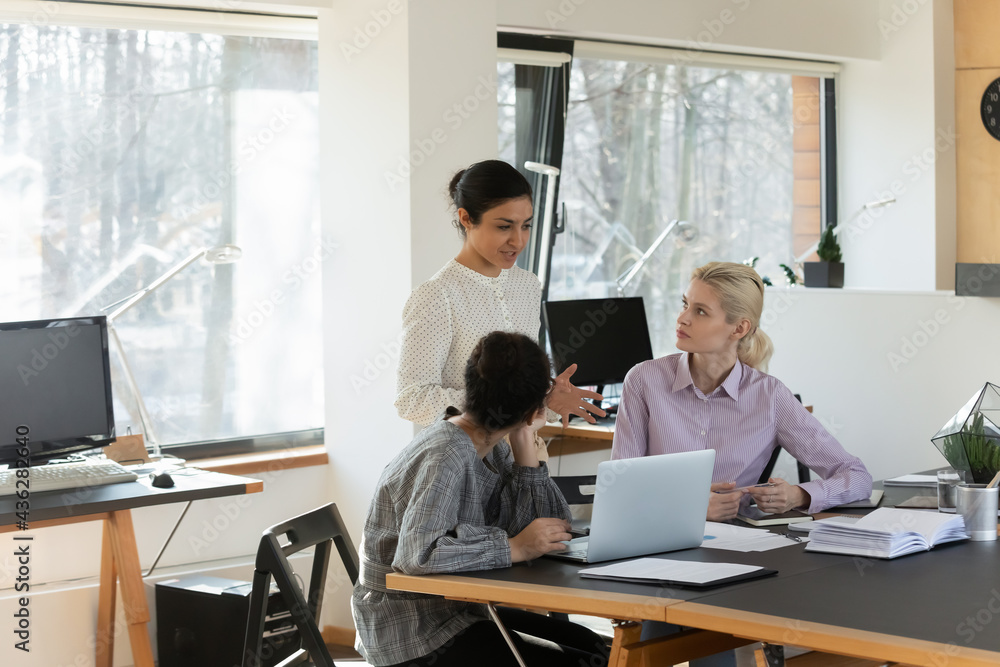 Diverse colleagues employees discussing strategy, working on project together, involved in briefing, brainstorming, two businesswomen interns listening to mentor coach, sitting at desk with laptop