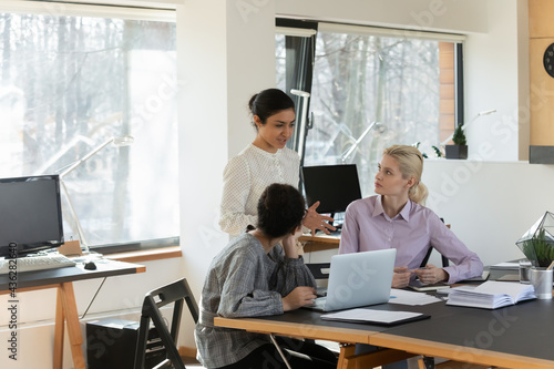 Diverse colleagues employees discussing strategy  working on project together  involved in briefing  brainstorming  two businesswomen interns listening to mentor coach  sitting at desk with laptop