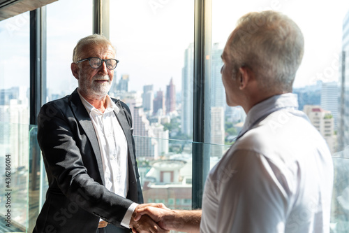 Two business men discussing their business together in a lobby of a hotel. They both wearing white shirt. The older one also wear black jacket. The scene outside is cityscape.