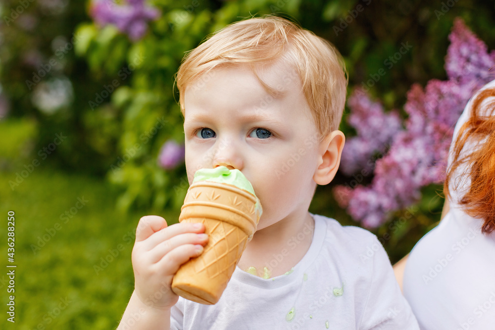 A little boy eats ice cream outdoors in a spring park on a blurry background of blooming lilacs.