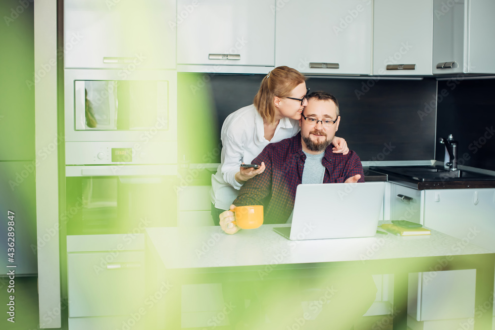Happy adult married couple using laptop in cozy kitchen interior. Woman in glasses embracing her husband. In the foreground leaves of houseplant, blurred focus. Family mutual understanding concept. Happy adult married couple using laptop in cozy kitchen interior. Woman in glasses embracing her husband. In the foreground leaves of houseplant, blurred focus. Family mutual understanding concept.