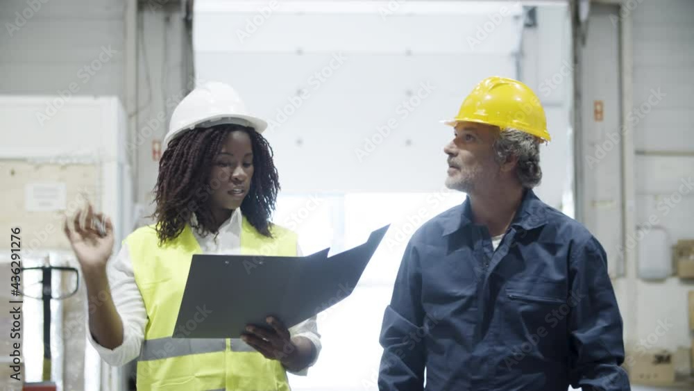 Focused workers walking through storehouse and talking. African American woman holding clipboard and reporting on goods received to her Caucasian coworker. Inventory, teamwork, warehouse concept Focused workers walking through storehouse and talking. African American woman holding clipboard and reporting on goods received to her Caucasian coworker. Inventory, teamwork, warehouse concept