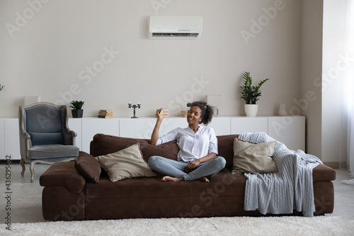 Full length relaxed smiling beautiful african ethnicity female homeowner sitting on comfortable sofa, turning on air conditioning system, breathing fresh air, enjoying peaceful moment indoors.