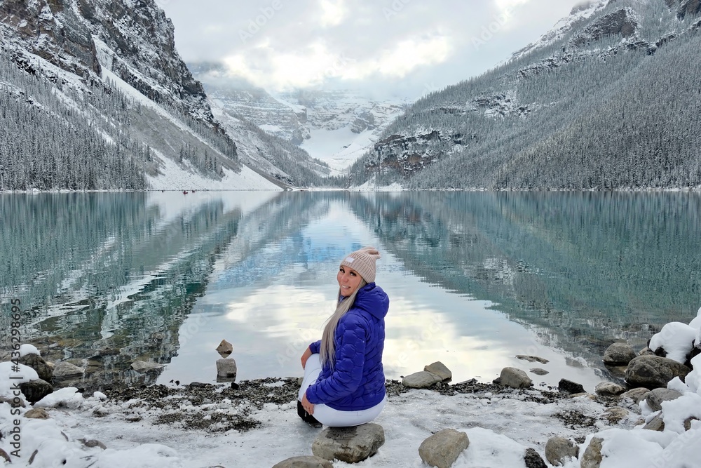 Fototapeta premium Attractive smiling woman in sport clothes on lake shore by snowy mountains. Early winter on Lake Louise in Banff National Park. Alberta. Canada
