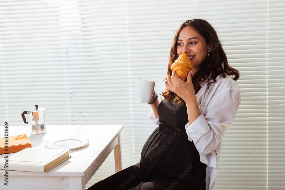 Happy pregnant young beautiful woman eating croissant during morning breakfast. Concept of pleasant morning and positive attitude during pregnancy Happy pregnant young beautiful woman eating croissant during morning breakfast. Concept of pleasant morning and positive attitude during pregnancy