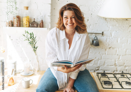 Gorgeous happy young woman plus size body positive in blue jeans and white shirt reading cooking book in the home kitchen