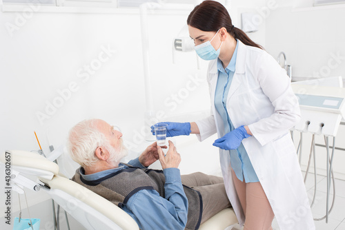 dentist giving glass of water to senior patient lying in dental chair.