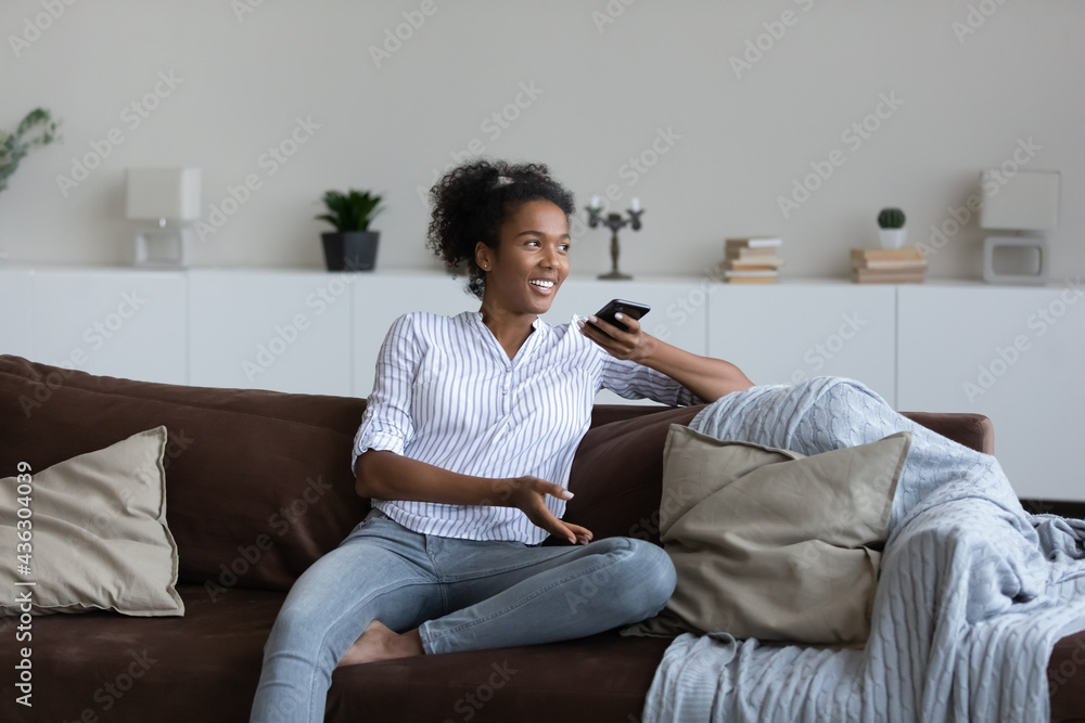 Happy young mixed race african woman resting on cozy couch, dictating audio message on cellphone, sending voicemail to friends, web surfing information online using virtual assistant application. Happy young mixed race african woman resting on cozy couch, dictating audio message on cellphone, sending voicemail to friends, web surfing information online using virtual assistant application.