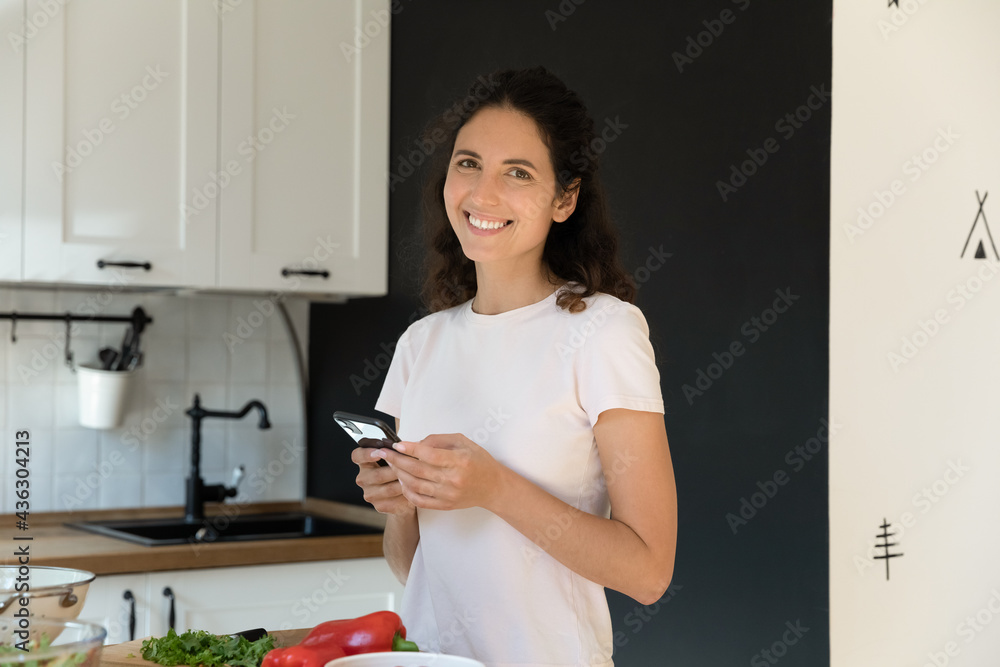 Joyful happy young hispanic housewife holding smartphone in hands, enjoying communicating in social network, distracted from cooking in kitchen. Smiling woman web surfing tasty recipe, preparing food. Joyful happy young hispanic housewife holding smartphone in hands, enjoying communicating in social network, distracted from cooking in kitchen. Smiling woman web surfing tasty recipe, preparing food.