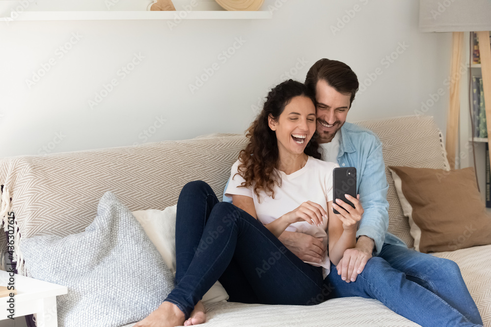 Happy bonding young family couple resting on couch, watching funny photos in social network, having fun entertaining playing mobile games together. Laughing spouses holding web camera video call. Happy bonding young family couple resting on couch, watching funny photos in social network, having fun entertaining playing mobile games together. Laughing spouses holding web camera video call.