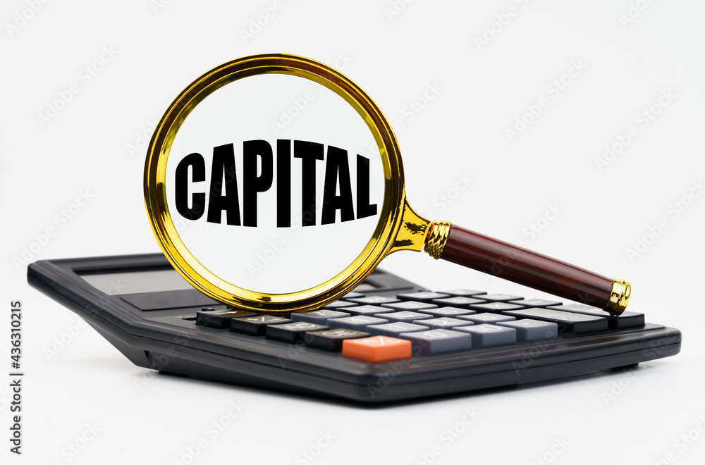 On a white background lies a calculator and a magnifying glass with the inscription - CAPITAL On a white background lies a calculator and a magnifying glass with the inscription - CAPITAL