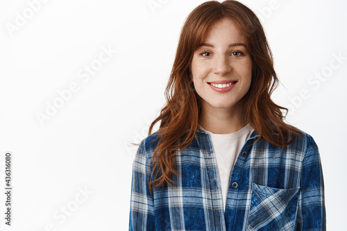Portrait of young redhead woman with natural healthy skin, freckles and white smile, looking happy and satisfied at camera, standing relaxed against white background