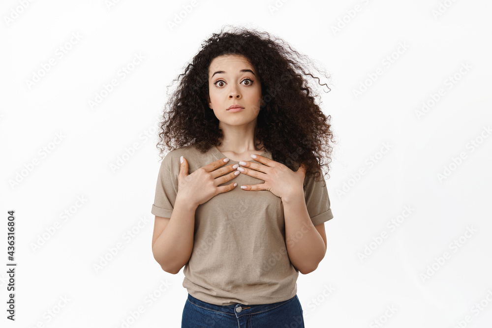 Surprised young woman looking startled, raising eyebrows in disbelief, holding hands on chest, being chosen unexpectedly, staring confused at camera, white background Surprised young woman looking startled, raising eyebrows in disbelief, holding hands on chest, being chosen unexpectedly, staring confused at camera, white background