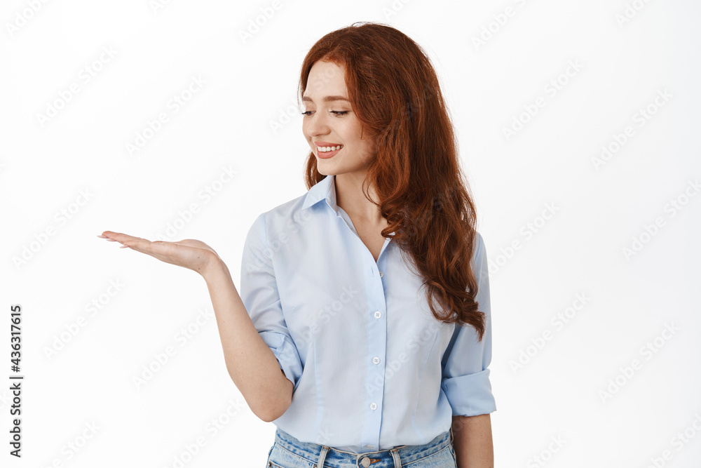 Cute redhead woman looking at open palm and smiling at product, holding item copy space in hand, standing in office blouse against white background Cute redhead woman looking at open palm and smiling at product, holding item copy space in hand, standing in office blouse against white background