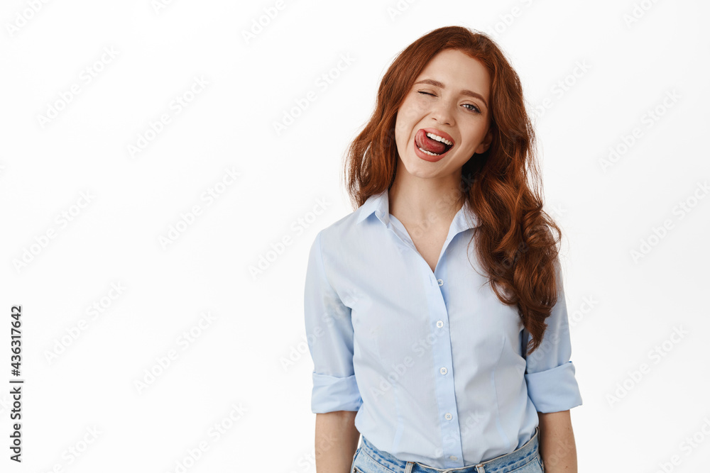 Cheerful candid redhead girl, smiling and showing tongue, winking at you carefree, standing relaxed and joyful against white background. Copy space Cheerful candid redhead girl, smiling and showing tongue, winking at you carefree, standing relaxed and joyful against white background. Copy space