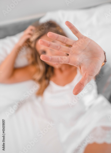 Portrait of an attractive young blond Caucasian woman in nightgown on the bed, putting her hand to the camera. Lifestyle in the hotel bed, overhead shot