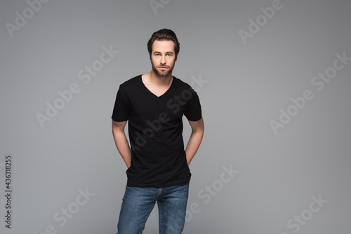 bearded man in black t-shirt and jeans posing with hands behind back isolated on grey.