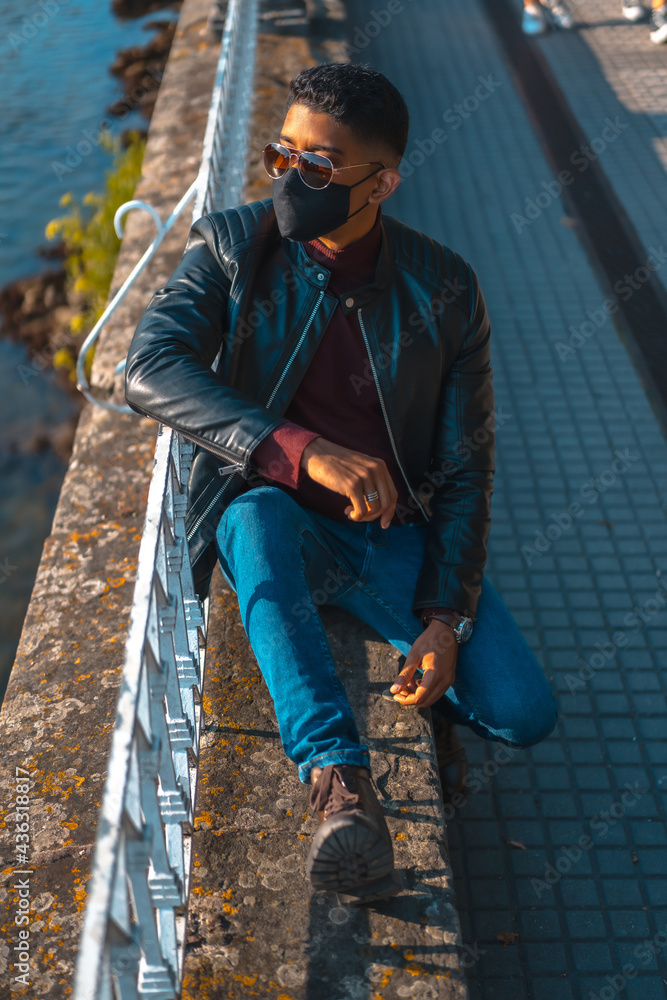 Fashion lifestyle, portrait of a young Latino in the river of the city. Jeans, leather jacket and brown shoes. In a pandemic with a mask