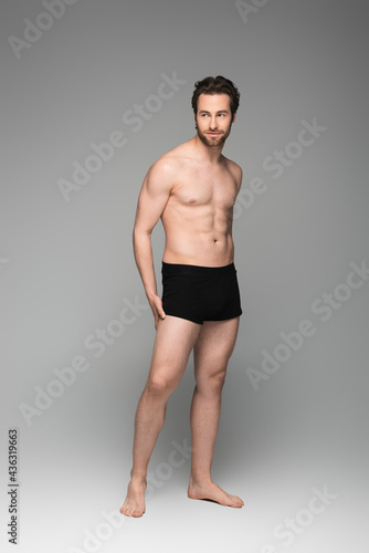 full length of bearded and shirtless man in black underwear posing on grey.
