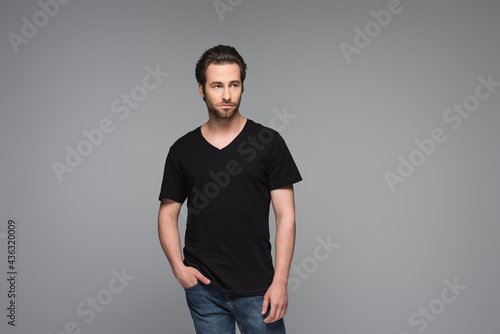 bearded man in black t-shirt and jeans posing with hand in pocket isolated on grey.