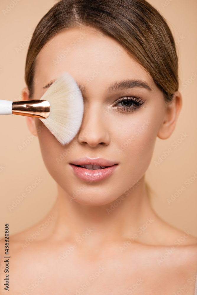 pretty woman with natural visage covering eye with cosmetic brush isolated on beige.