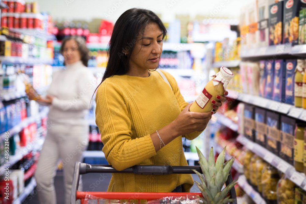 Pensive latina buying groceries at store, walking with shopping cart among shelves and choosing products