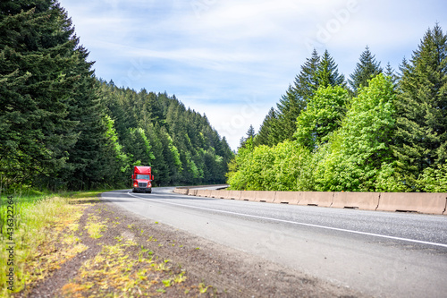 Bright red big rig semi truck with semi trailer in a hurry for timely delivery of goods running on the green forest road